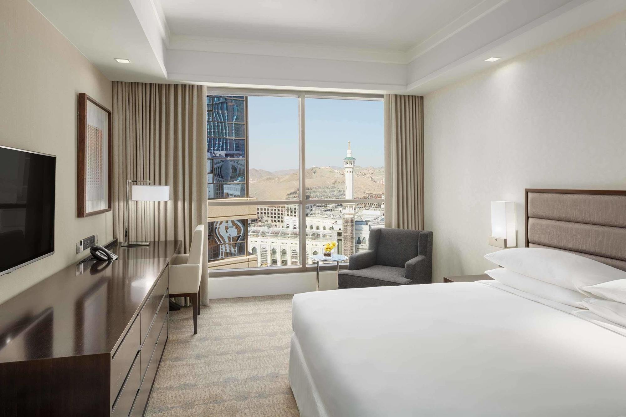 Jabal Omar Hyatt Regency Makkah Hotel Mecca Exterior photo The photo shows a modern hotel room featuring a large window that provides a view of an outdoor landscape, likely part of a city or landmark. The room includes a bed with white linens, a side table with a lamp, and a small seating area with a chair a