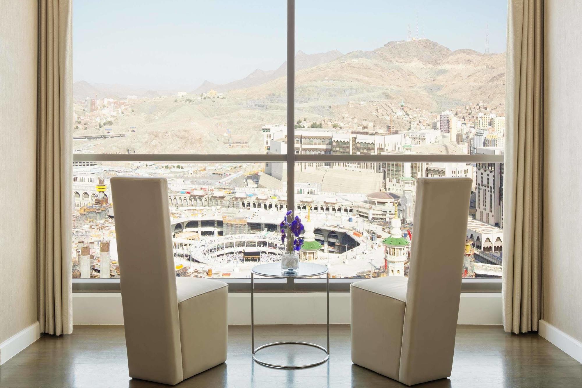 Jabal Omar Hyatt Regency Makkah Hotel Mecca Exterior photo The photo shows a modern indoor setting with two tall, white chairs positioned facing a large window. Through the window, there is a panoramic view of a cityscape that includes mountainous terrain and buildings, suggesting it may be a notable locatio