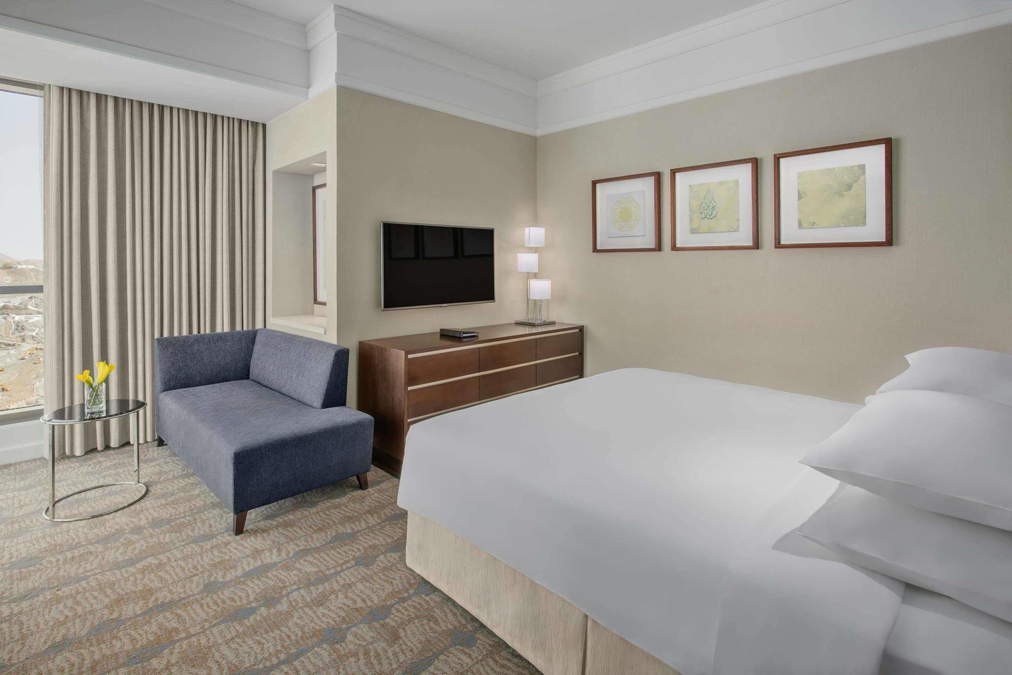 Jabal Omar Hyatt Regency Makkah Hotel Mecca Exterior photo The photo shows a modern hotel room featuring a neatly made bed with white linens, a small blue couch to the side, and a wooden dresser with a television mounted on the wall above it. There are two framed pieces of art on the wall near the bed, and t