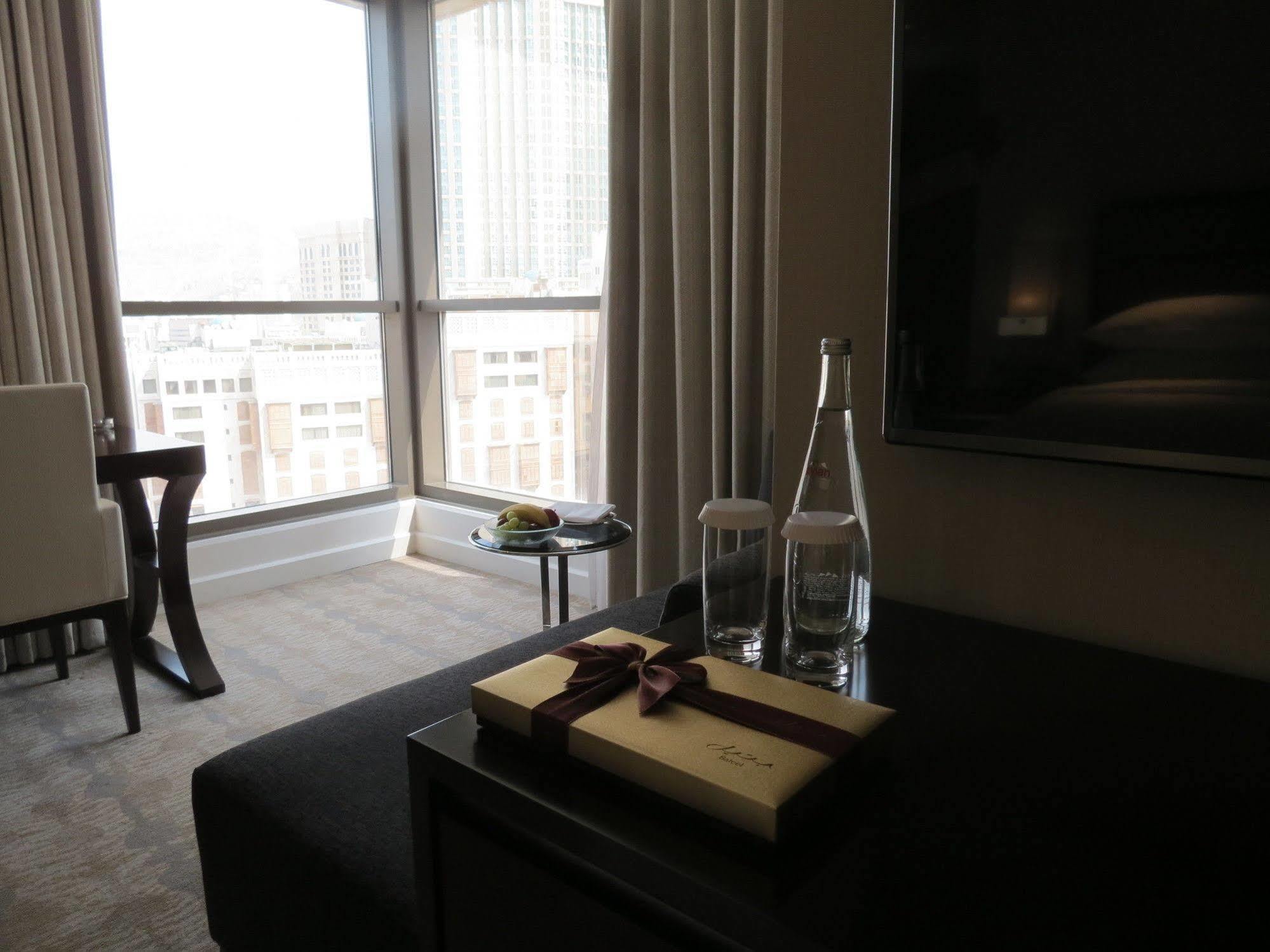 Jabal Omar Hyatt Regency Makkah Hotel Mecca Exterior photo The photo shows a modern hotel room with large windows letting in natural light. In the foreground, there is a table with a decorative box tied with a ribbon, two clear glass water bottles, and a couple of glasses. In the background, there's a comfor